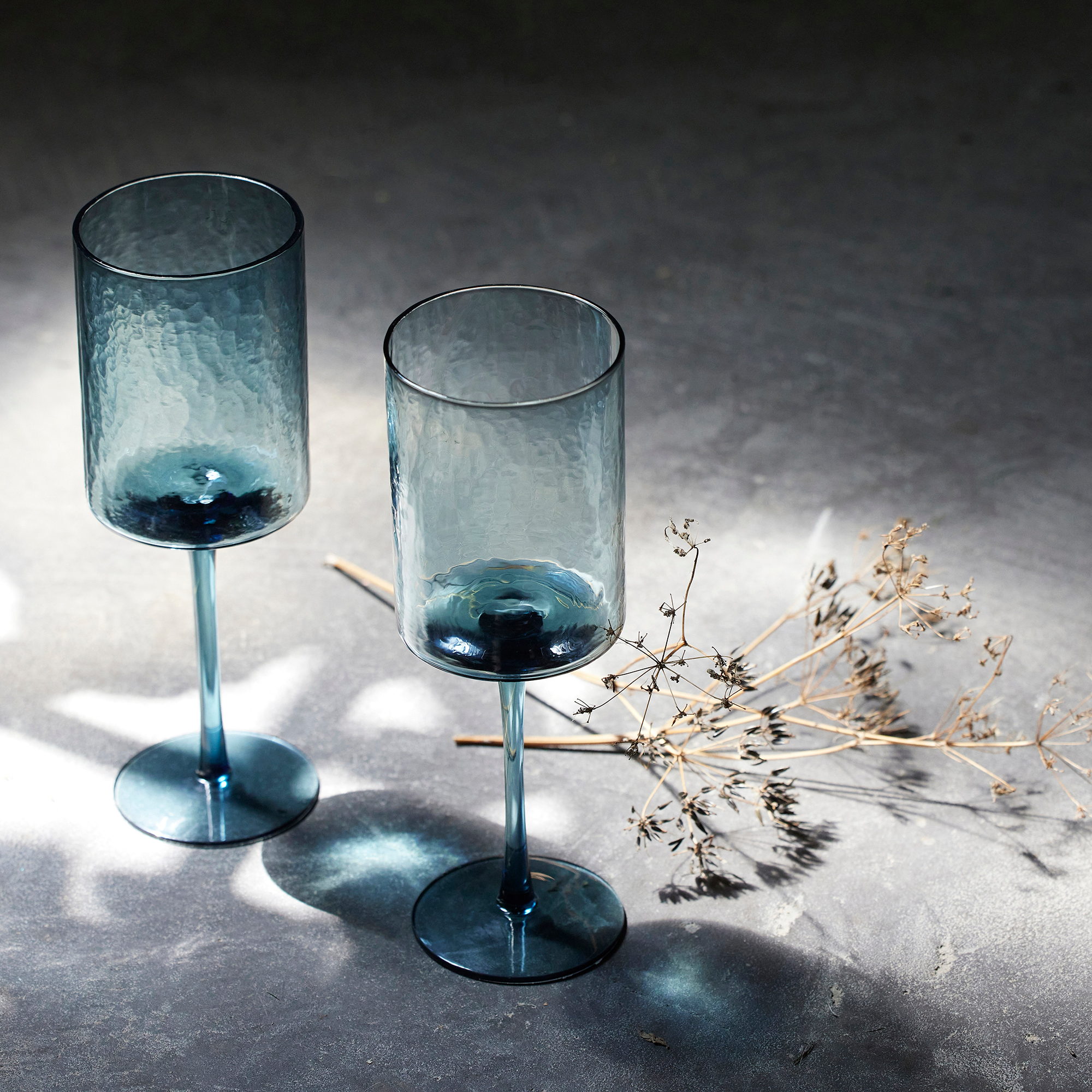 Deux verres à pied bleus et une ombelle séchée sur un plan de travail clair, lumière rasante